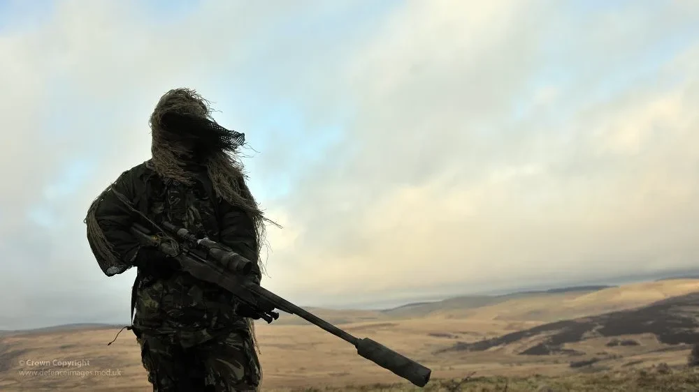 Snajper w kombinezonie ghillie z karabinem wyborowym z tłumikiem obserwuje otwarty teren z grzbietu wzgórza